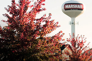 A watertower for the Village of Marshall is shown in the image. Itis white with Marshall in white letters against a red background. A tree with red leaves is in the left of the frame.