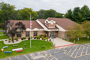 This image shows the village hall for Cottage Grove. There is a flagpole outside the building, and a parking lot is in the foreground.