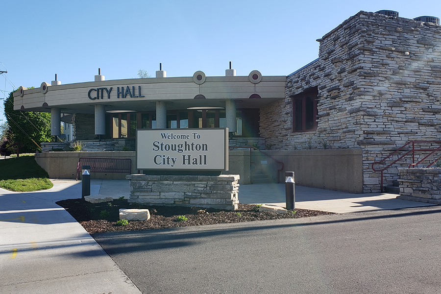 Stoughton City Hall is shown in this image. A sign out front reads Stoughton City Hall, and City Hall is spelled out in block letters on the building.