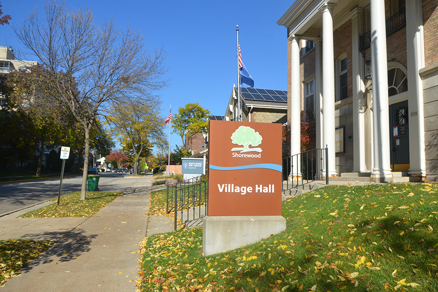 A sign for the Shorewood Village Hall is shown in this image. A building with columns is to the right and a flag pole is in the background.
