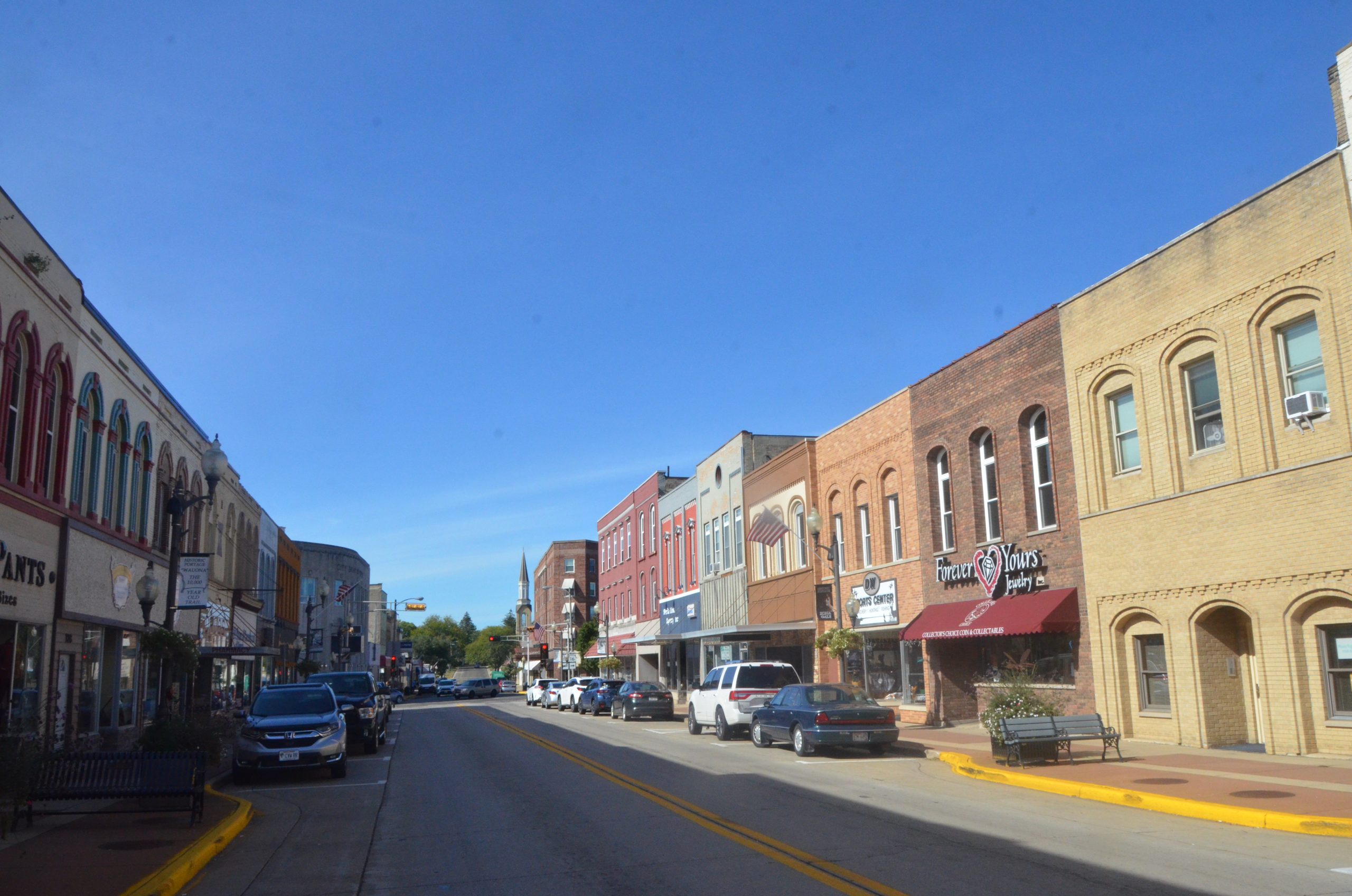 A downtown streetscape in the City of Portage. Buildings line either side of a road with parked cars alongside it.